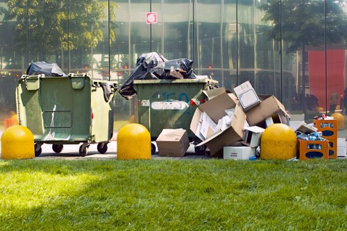 A row of three large wheelie bins positioned on a grassy area in front of a building with glass windows reflects trees and sky. The first green bin on the left has a black plastic bag partially covering the top, while the middle green bin is empty with a closed lid. The third bin, also green, is partially obscured by overflowing cardboard boxes and plastic containers, some of which are flattened or damaged, spilling onto the ground. The cardboard boxes vary in size, with some showing visible tape or labels. Bright yellow plastic bollards are situated in front of the bins to prevent vehicle impact. The environment suggests an outdoor commercial or industrial setting typical for rubbish disposal, with natural daylight illuminating the scene and a street sign visible in the background. The arrangement indicates waste collection or clearance, consistent with services provided by Commercial Waste Greenhithe for efficient rubbish removal in the local area.