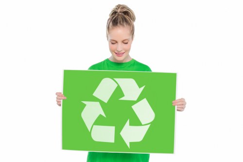 A young woman with light brown hair tied back in a ponytail, wearing a bright green long-sleeved shirt, is holding a large rectangular green sign featuring a white recycling symbol composed of three chasing arrows. She is smiling and standing against a plain white background. The sign and her attire suggest an emphasis on environmental awareness and waste recycling efforts, aligning with environmentally conscious rubbish removal services such as those provided by Commercial Waste Greenhithe. The image conveys a positive message about recycling and waste minimisation, which is relevant to waste management initiatives in the Greenhithe area. The woman appears to be promoting sustainable waste disposal practices, supporting local efforts to encourage recycling and eco-friendly rubbish management solutions, in a professional and approachable manner.