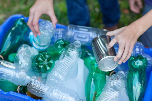 A close-up view of a person's hands sorting through a collection of discarded plastic bottles and cans, including clear PET bottles and green plastic bottles with black caps, placed in a blue recycling bin. The background shows a person wearing blue jeans and a dark shirt, standing outdoors on a grassy area, indicating a site for waste collection or disposal. The scene is well-lit with natural daylight, highlighting the textures and colours of the plastic materials, which are arranged on the top of the bin, ready for collection or recycling. This image relates to rubbish removal services in Greenhithe, where Commercial Waste Greenhithe manages the collection and disposal of various types of waste materials, including plastics, from commercial establishments or residential areas near the postcode area.