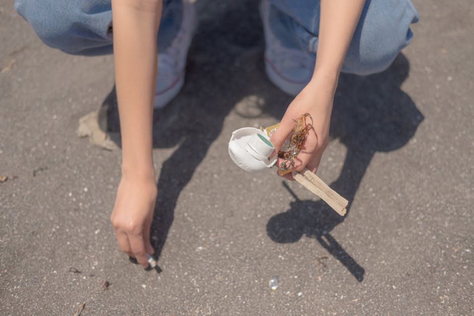 A person crouching on an outdoor paved surface, holding a small collection of debris including a white plastic cup, a piece of scrap wood, and tangled string or twine, with their other hand resting on